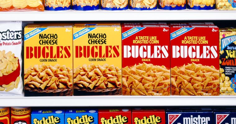 Image of two yellow boxes of Nacho Cheese Bugles aside two red boxes of original Bugles on a store shelf next to other snack products for sale.